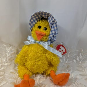 May include: A plush yellow duck toy with orange feet and beak, wearing a blue and white checkered bonnet and a light blue ribbon. The toy has black eyes and a red tag.