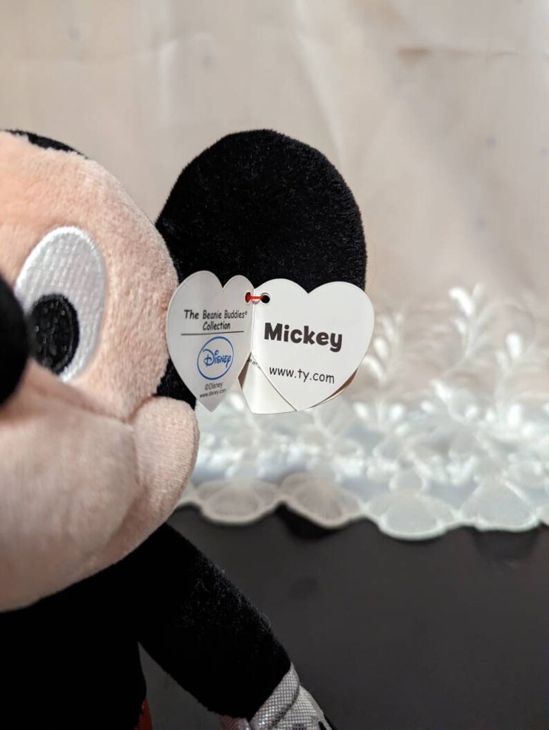 May include: A plush Mickey Mouse toy from the Beanie Buddies Collection. The tag is a heart shape with the text "Mickey" and "www.ty.com". The tag also has a Disney logo.