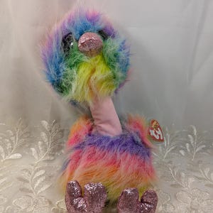 May include: A colorful plush ostrich toy with rainbow-colored feathers. The toy has large, glittery eyes and a glittery pink beak. The ostrich has pink heart-shaped feet and a pink neck. A red and white tag is attached to the toy.