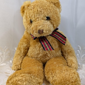 May include: A plush teddy bear with golden brown fur. The bear has a brown nose, black eyes, and a colorful striped ribbon tied around its neck. The bear is sitting on a white surface.