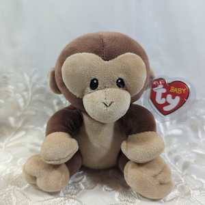 May include: A brown and tan plush monkey toy with a red "Ty" tag that says "Baby" on it. The monkey is sitting on a white lace surface.