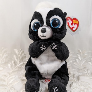 May include: A soft toy skunk, predominantly black and white, with a white stripe and large blue eyes. It has a red heart-shaped tag attached. The toy is designed to be cuddly.