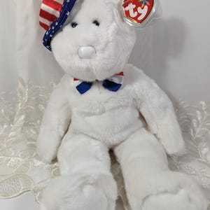 May include: White plush teddy bear wearing a red, white, and blue patriotic hat and bow tie. The bear has a Ty tag attached to its ear.