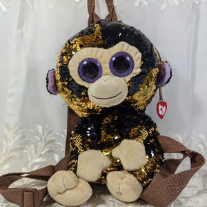 Peut inclure: Un sac à dos singe en sequins dorés et noirs avec de grands yeux violets. Le sac à dos a une sangle réglable marron et une étiquette Ty.