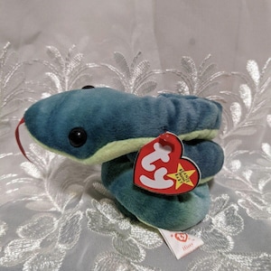 May include: A teal and green plush snake toy with large black eyes and a red tongue. It has a red heart-shaped tag that reads "Beanie Babies" and a smaller tag that says "Hissy".