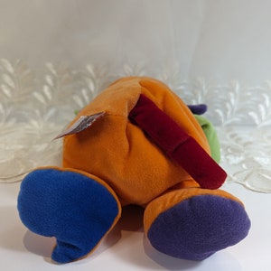 Pode incluir: Um brinquedo de pel&uacute;cia multicolorido com a forma de um animal e uma cauda longa vermelha. O brinquedo est&aacute; deitado de costas com as patas no ar. O brinquedo &eacute; feito de tecido macio e felpudo e tem uma variedade de cores, incluindo laranja, azul, verde e roxo.