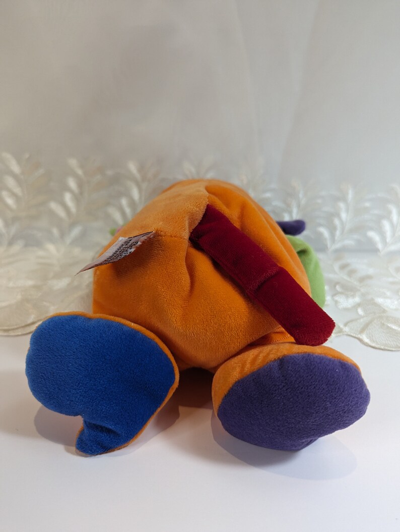 Pode incluir: Um brinquedo de pel&uacute;cia multicolorido com a forma de um animal e uma cauda longa vermelha. O brinquedo est&aacute; deitado de costas com as patas no ar. O brinquedo &eacute; feito de tecido macio e felpudo e tem uma variedade de cores, incluindo laranja, azul, verde e roxo.