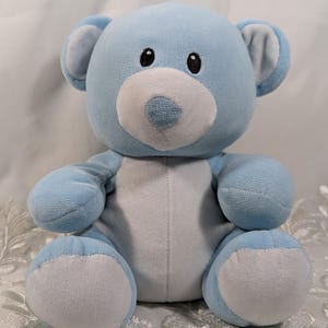 May include: A plush teddy bear in light blue and white. The bear has a light blue body, arms, and legs, with white accents on the snout, belly, and the soles of its feet. It has black eyes and light blue ears.