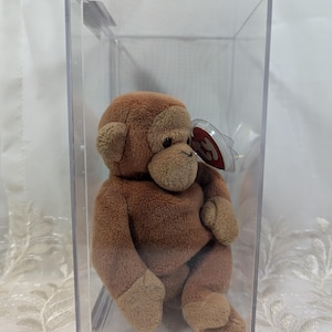 May include: A brown beanie baby monkey in a clear plastic display case. The monkey is sitting with its legs crossed and its arms folded. The beanie baby has a red tag with the words "Ty" and "Beanie Babies" on it.