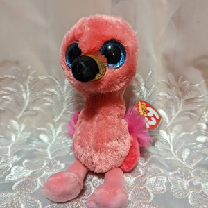 Può includere: Flamingo di peluche rosa con grandi occhi blu e un becco nero. Il fenicottero è seduto con le gambe incrociate e ha un'etichetta Beanie Boo rosa e bianca attaccata all'ala.