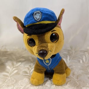 May include: A plush toy of a cartoon dog, wearing a blue police hat and vest. The dog has large, round eyes, brown and tan fur, and a black nose. The hat features a badge, and the vest has a star.