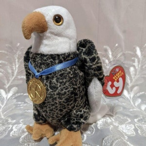 Peut inclure: Une peluche d'aigle à tête blanche, bec marron et corps noir et gris moucheté. Il porte un ruban bleu avec un médaillon doré. Une étiquette rouge avec du texte blanc est attachée.