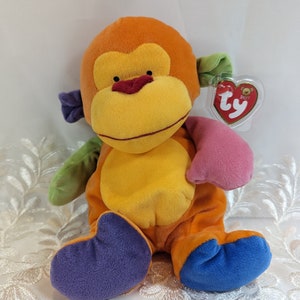 Pode incluir: Um brinquedo de macaco de pel&uacute;cia multicolorido com uma etiqueta Ty Beanie Baby. O macaco est&aacute; sentado com as pernas cruzadas e tem um esquema de cores roxo, verde, laranja e azul.