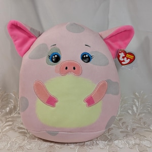 May include: A plush pig toy featuring a light pink body with grey spots, a pale yellow belly patch, and bright pink inner ears. The pig has embroidered blue eyes with long lashes and a small pink snout. A heart-shaped Ty Squishy Beanies tag is attached.