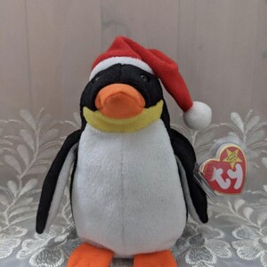 May include: A black and white penguin wearing a red Santa hat with a white pom-pom. The penguin has a yellow beak and orange feet. The penguin is holding a red Ty Beanie Baby tag with a white star and the word "Ty" in white.