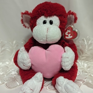 May include: A red plush monkey toy with white details on its face, hands, and feet, clutching a pink heart. The monkey has a tag attached. The toy is on a white surface.