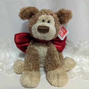 May include: A brown and cream plush dog toy with a red satin bow. The toy has a brown nose and black eyes. The tag on the toy reads "Gotta Getta Gund".
