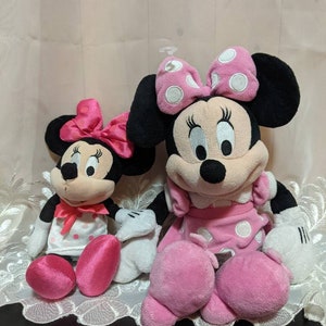 May include: Two plush Minnie Mouse toys, one larger than the other, both wearing pink dresses with white polka dots and pink bows. The larger Minnie Mouse has black ears and a white face with black eyes and a black nose. The smaller Minnie Mouse has pink ears and a white face with black eyes and a black nose.