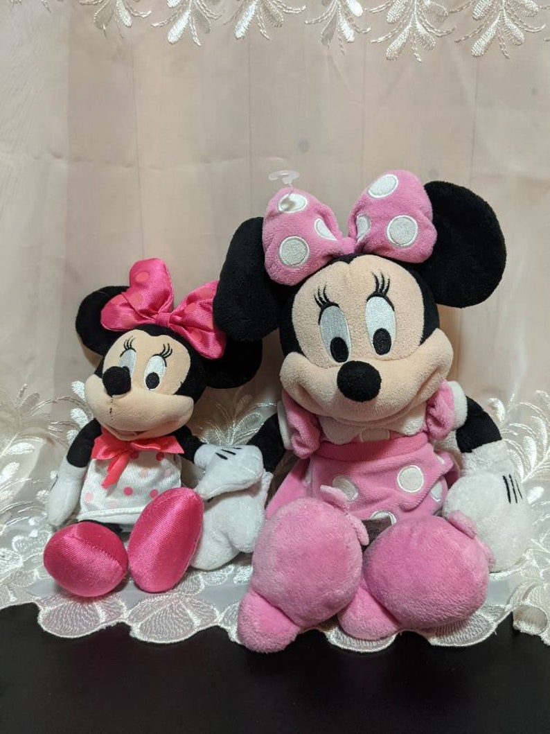 May include: Two plush Minnie Mouse toys, one larger than the other, both wearing pink dresses with white polka dots and pink bows. The larger Minnie Mouse has black ears and a white face with black eyes and a black nose. The smaller Minnie Mouse has pink ears and a white face with black eyes and a black nose.