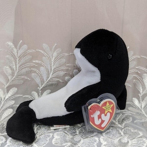 May include: Black and white plush orca whale toy with a Ty Beanie Baby tag. The tag is red with white text and a gold star.