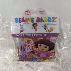 May include: A package of 12 colorful rubber band bracelets featuring Dora the Explorer. The package is purple and pink with the words "Beanie Bandz" and "Dora Collection".