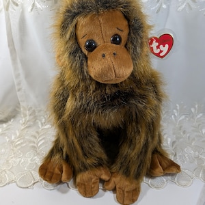 May include: A plush orangutan toy with brown and tan fur, large dark eyes, and a soft, textured face. The toy has a Ty heart-shaped tag attached. The orangutan is sitting on a white surface.