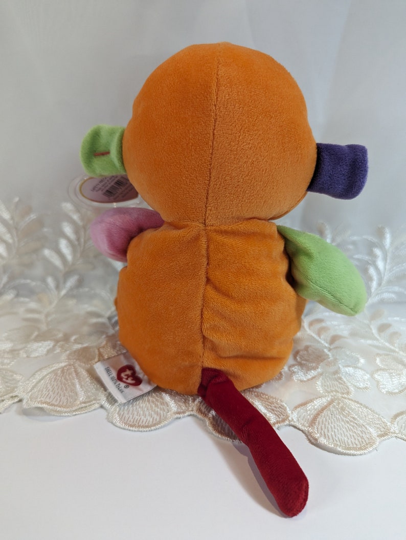 Pode incluir: Um macaco de pel&uacute;cia multicolorido com uma cauda vermelha. O macaco est&aacute; sentado em uma renda branca. O macaco tem um design de patchwork verde, rosa e roxo. O macaco tem uma etiqueta que diz "Ty Beanie Babies".