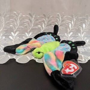 May include: A colorful butterfly Beanie Baby with black wings, a green head, and a rainbow-colored body. The Beanie Baby has a red and white Ty tag attached to it.