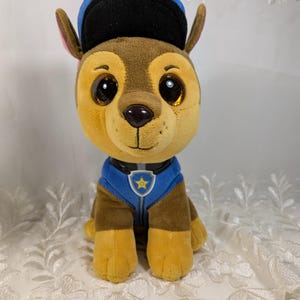 May include: A plush toy of a cartoon dog, wearing a blue police hat and uniform. The dog has large, glittery eyes, brown fur, and yellow paws. The uniform features a star emblem. The toy is sitting upright.