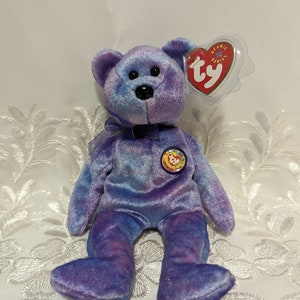 Può includere: Un orsacchiotto Beanie Baby di peluche nei toni del viola e del blu. L'orso ha un naso nero e un piccolo fiocco intorno al collo. Ha un'etichetta a forma di cuore con la scritta "Beanie Babies" e un'etichetta circolare con la scritta "Official Club".