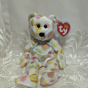May include: White plush Easter-themed beanie baby bear with a blue ribbon around its neck. The bear is covered in colourful Easter egg patterns. The bear has a black nose and black eyes. The bear is sitting on a white lace surface. The bear has a Ty Beanie Babies tag attached to its right ear.