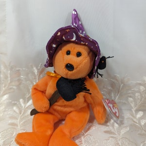 Può includere: Orso di peluche arancione con un cappello da strega viola con stelle e un design a mezzaluna. L'orso tiene una scopa nera e ha un ragno nero sul cappello. L'orso ha un'etichetta Ty attaccata al braccio.