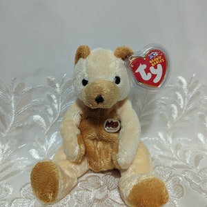 Peut inclure: Un ourson en peluche Ty Beanie Baby de couleur beige et marron nommé "Happy" avec une étiquette rouge "Ty". L'étiquette indique "Beanie Babies" et a la forme d'un cœur. L'ourson est assis sur une surface en dentelle blanche.