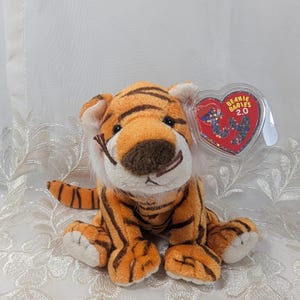 Könnte beinhalten: Orange-schwarz gestreiftes Plüschtier Beanie Baby Tiger mit weißem Bauch und weißen Pfoten. Der Tiger sitzt mit leicht geneigtem Kopf. Das Spielzeug hat ein rotes herzförmiges Etikett mit der Aufschrift "Beanie Babies 2.0" in weißer Schrift.