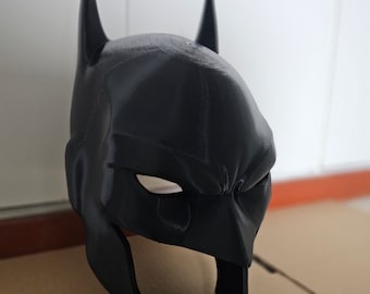 Könnte beinhalten: Eine schwarze Batman-Maske mit spitzen Ohren und Augenöffnungen. Die Maske besteht aus einem glatten, festen Material und ist so konzipiert, dass sie den gesamten Kopf bedeckt. Es ist eine Nachbildung der Kapuze des berühmten Superhelden.