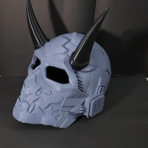 Puede incluir: Un casco integral de color gris claro con un diseño mecánico complejo y angular con una estructura similar a un cráneo. Dos cuernos puntiagudos, lisos y negros sobresalen de la parte superior del casco, que también incluye paneles laterales rectangulares.