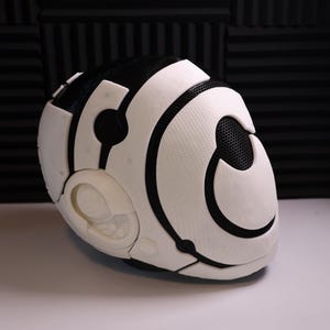 May include: A futuristic-looking helmet in white and black, with a mesh section and geometric shapes. The helmet is on a white surface, with a black sound-dampening panel in the background.