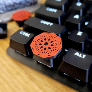 Puede incluir: Un tapón de tecla rojo impreso en 3D con un diseño geométrico, colocado en un teclado negro.