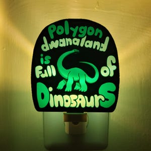 May include: A green night light with a black background and the text "Polygon Dwanaland is full of Dinosaurs". The night light features a green dinosaur silhouette.