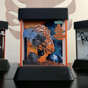 May include: A 3D-printed display featuring a layered design with the text "Ice Death Planets Lungs Mushrooms and Lava." The artwork has orange, blue, and black elements, set against a starry background. The display is housed in a black and orange frame.