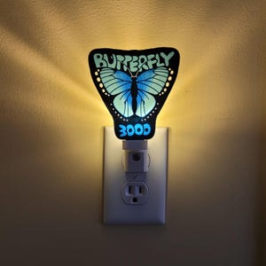 Può includere: Una luce notturna a forma di farfalla blu e verde con le parole "BUTTERFLY" e "3000" stampate su di essa. La luce notturna è collegata a una presa di corrente standard.