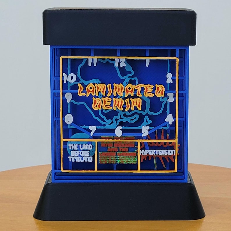 May include: A blue and black 3D printed clock with a clock face that says "Laminated Denim". The clock face also has a map-like design with numbers around the edge. The clock is on a black base.