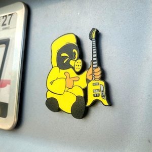 Puede incluir: Una figura de dibujos animados amarilla con un traje de protección que sostiene una guitarra eléctrica amarilla. La figura tiene una cara negra con una máscara de gas y señala con un dedo. La guitarra tiene detalles en blanco y negro.