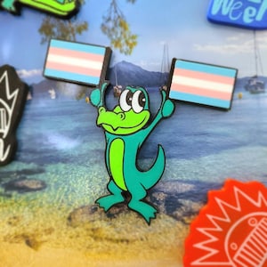 May include: A teal and green cartoon alligator holds two transgender pride flags. The flags are light blue, pink, and white. The alligator has a white belly and a black outline. Other colorful stickers are in the background.