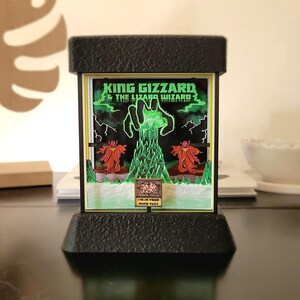 May include: A black, rectangular light-up display featuring the text "KING GIZZARD & THE LIZARD WIZARD" in green. The display shows a green hand reaching up from a green structure, with two red figures on either side. The bottom reads "I'M IN YOUR MIND FUZZ".