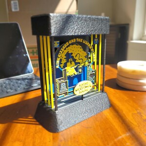 May include: A black metal display stand with a yellow and blue graphic of a cartoon character with the text "King Gizzard and the Lizard Wizard" and "Flying Microtonal Banana".