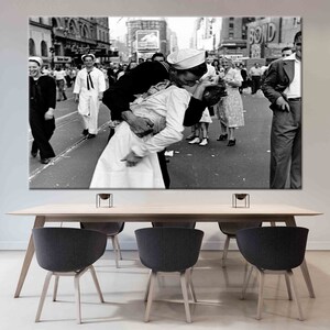 Può includere: Una fotografia in bianco e nero di un marinaio che bacia una donna a Times Square, New York City. La foto è scattata da un basso angolo, mostrando la coppia in primo piano e una folla di persone sullo sfondo. Il marinaio indossa un cappello bianco e un'uniforme, e la donna indossa un vestito bianco. La foto è un'immagine famosa della fine della seconda guerra mondiale.