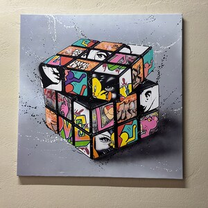 May include: A colorful, abstract painting of a Rubik's Cube with various images and patterns on each side. The cube is painted in a 3D style on a gray background.