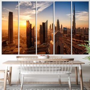 May include: A five-panel canvas print depicting a cityscape at sunset. The image shows tall, modern skyscrapers in shades of brown and gold, with the sun setting in the background. The print is displayed above a wooden dining table with white chairs.