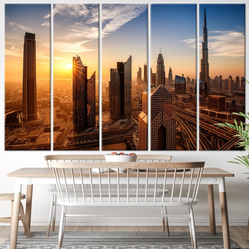 May include: A five-panel canvas print depicting a cityscape at sunset. The image shows tall, modern skyscrapers in shades of brown and gold, with the sun setting in the background. The print is displayed above a wooden dining table with white chairs.